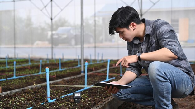 Slimme landbouwer die digitale tablettablet gebruikt die de groei van planten in de kas onderzoekt.