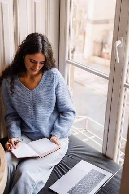 Slimme meid met laptop maakt aantekeningen in notitieboekje bij groot raam thuis Geconcentreerde jonge vrouw studeert online les met laptop op de bank bij groot raam