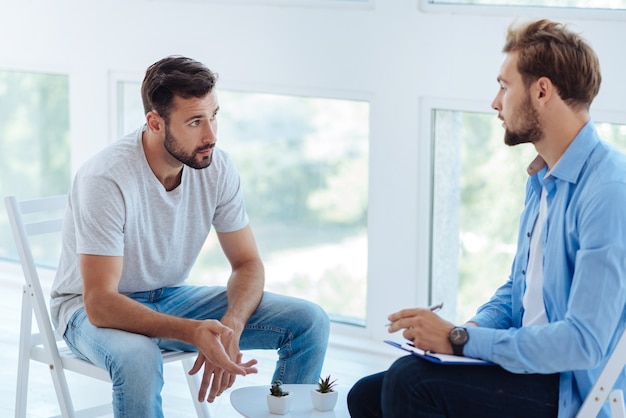 Slimme professionele mannelijke therapeut die naar zijn patiënt kijkt en aantekeningen maakt terwijl hij over zijn problemen luistert