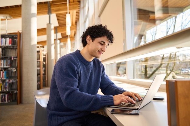 Slimme Spaanse student die aan een bureau zit en een laptop gebruikt om een project voor te bereiden op de bibliotheekcampus