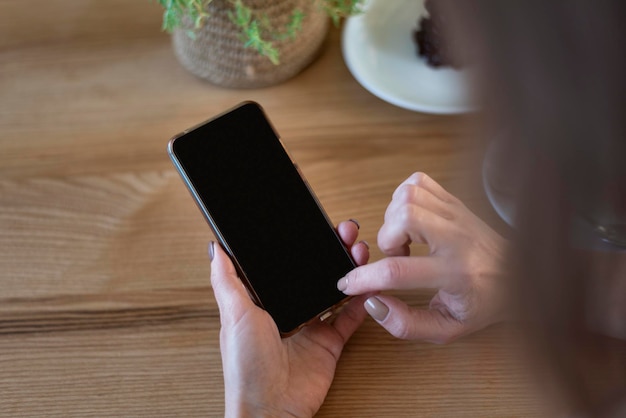 Slimme telefoon in handen van de vrouw tegen op houten tafel achtergrond Smartphone met leeg zwart scherm Kopieer ruimte