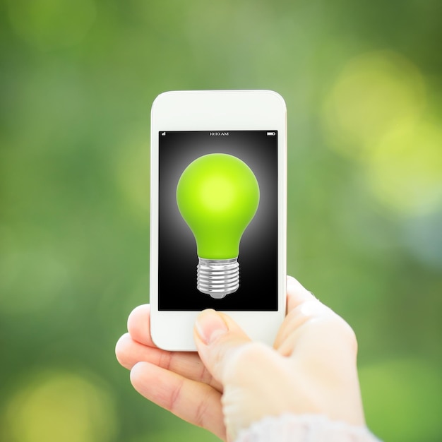 Slimme telefoon met in hand gloeilamp tegen groene de lenteachtergrond Ecologieconcept