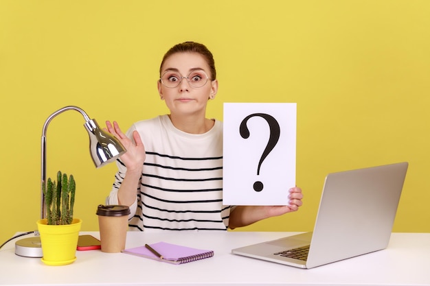 Foto slimme vrouw kantoormedewerker zittend op de werkplek met vraagteken met twijfelachtige uitdrukking