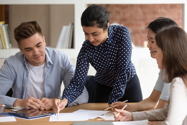 Slimme vrouwelijke indiase teamleider houdt groepsbijeenkomst met studenten legt uit studiemateriaal tekenen op papier multiculturele jongeren verzamelen zich aan tafel bespreken project of opdracht Teamwerkconcept