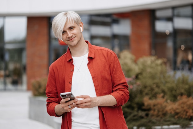 Slimme zelfverzekerde tiener met zijn telefoon buiten. Portret van blonde jonge hipster met zijn telefoon.