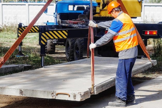 Slinger lays concrete slab on construction site on summer day Worker in protective vest and construction helmet supervises laying of base on construction site