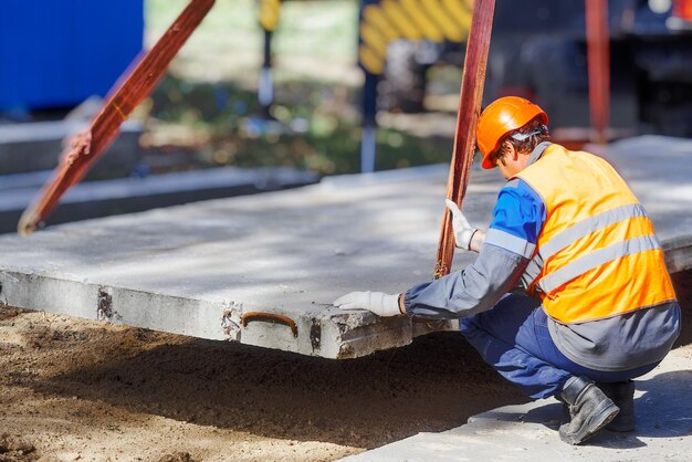 Slinger lays concrete slab on construction site on summer day Worker in protective vest and construction helmet supervises laying of base on construction site