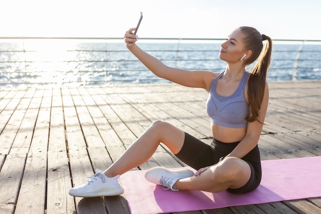 Slm fitte vrouw in sportkleding maakt selfie op een smartphone terwijl ze bij zonsopgang op de mat op het strand zit
