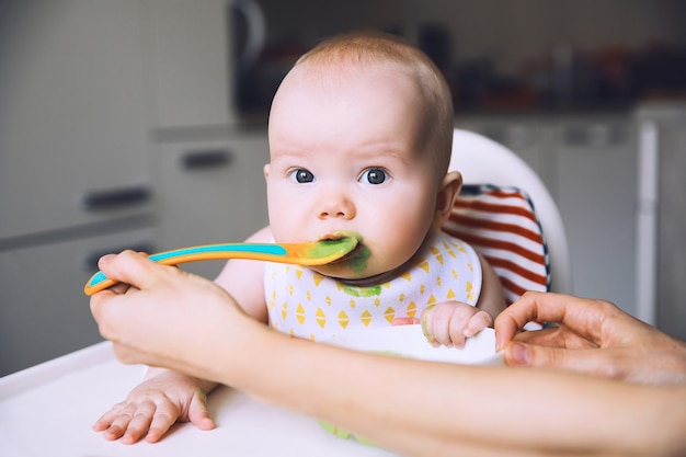 Slordige lachende baby voeden die met een lepel in de kinderstoel eet Baby's eerste vaste voedsel