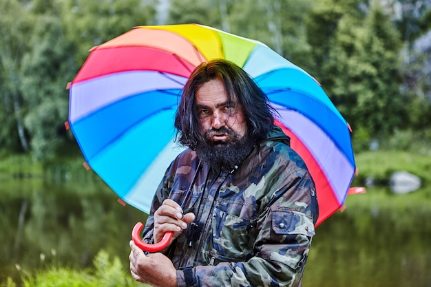 Slordige ruige man, 53 jaar oud, met een geschilderde baard en pruilende lippen, verbergt zich voor regen onder een regenboogparaplu.