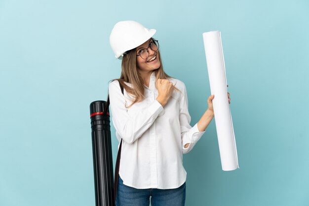 Slovak architect girl holding blueprints isolated on blue wall celebrating a victory