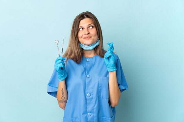 Dentista slovacco che tiene gli strumenti isolati su sfondo blu con le dita incrociate e augurando il meglio