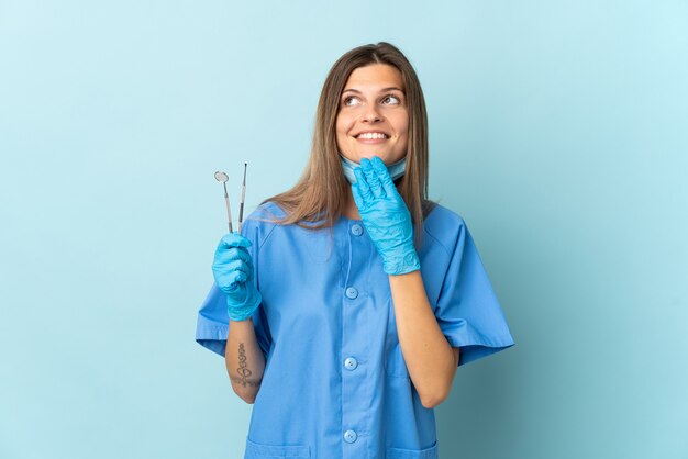 Slovak dentist holding tools isolated on blue wall looking up while smiling