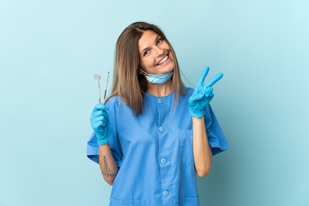 Slovak dentist holding tools isolated on blue wall smiling and showing victory sign