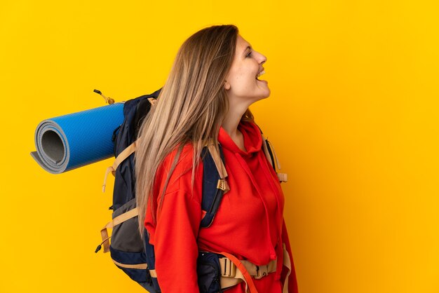 Slovak mountaineer woman with a big backpack isolated on yellow background laughing in lateral position