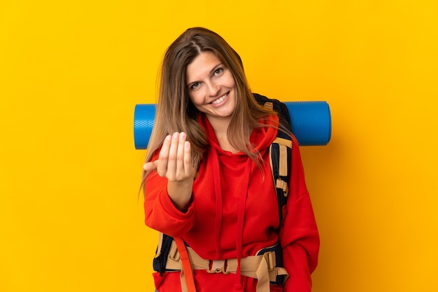 Slovak mountaineer woman with a big backpack isolated on yellow wall inviting to come with hand. Happy that you came