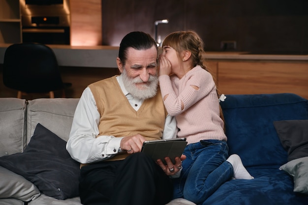 Slow motion of cute joyful 12-aged girl with funny pigtails which whispering on granddad's ear her secret while they sitting together on the sofa at home