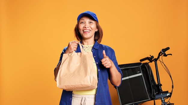 Slow motion portrait shot of bored food delivery bike rider waiting for customer, showing exultant approving gesturing. Asian courier isolated over studio background providing meal order to client