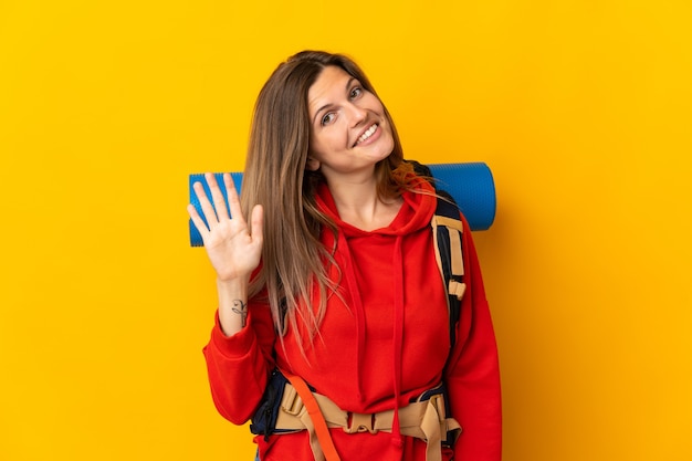 Slowaakse bergbeklimmervrouw met een grote rugzak die op gele achtergrond wordt geïsoleerd die met hand met gelukkige uitdrukking salueert