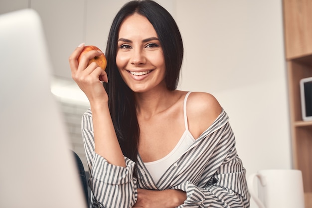 Sluit me aan bij mij. Blij brunette meisje dat op afstand werkt, achter haar laptop in de keuken zit