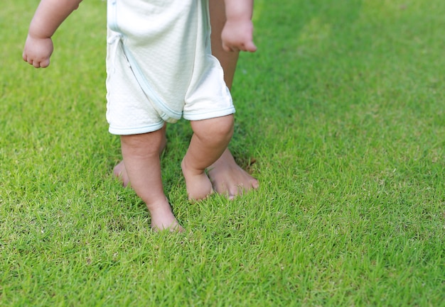 Sluit omhoog de voeten die van de zuigelingsbaby om met zijn moeder op het groene gras leren te lopen