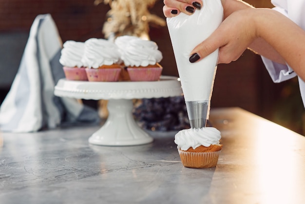 Foto sluit omhoog handen van de vrouwelijke chef-kok met suikergoedzak die room op cupcakes drukken.