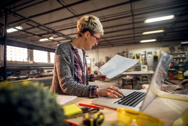 Foto sluit omhoog mening van charmante glimlachende gemotiveerde korte haar vrouwelijke ingenieur met oogglazen die met blauwdrukken en laptop in de workshop werken