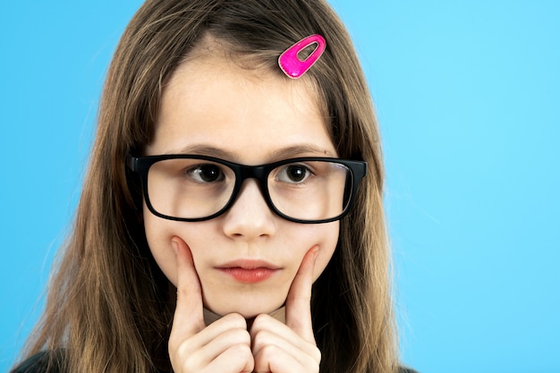 Sluit omhoog portret van een kindschoolmeisje die kijkend glazen dragen die hand houden aan haar gezicht denkend over iets geïsoleerd op blauwe muur