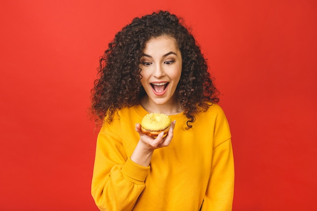 Sluit omhoog portret van een tevreden vrij jong meisje dat donuts eet dat over rode achtergrond wordt geïsoleerd.