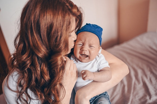 Sluit omhoog portret van gelukkig jong mamma die haar schreeuwende baby in jeans en witte t-shirt en GLB houden. Jonge gelukkige familie, moeder spelen met schattige emotionele kleine pasgeboren kind-zoon in de slaapkamer.