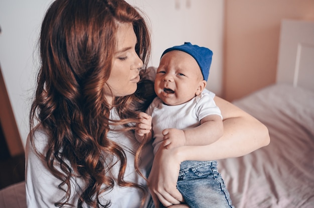 Sluit omhoog portret van gelukkig jong mamma die haar schreeuwende baby in jeans en witte t-shirt en GLB houden. Jonge gelukkige familie, moeder spelen met schattige emotionele kleine pasgeboren kind-zoon in de slaapkamer.