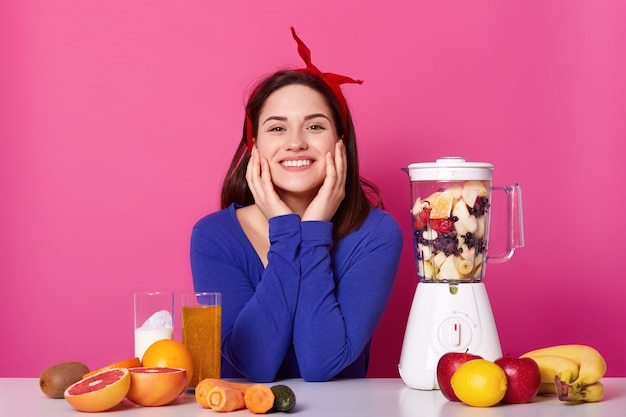 Sluit omhoog portret van leuke glimlachende jonge vrouw met gelukkige uitdrukking, houdt haar handen op wangen, gefotografeerd in fotostudio geïsoleerd op roze met veel fruit en blendrer.