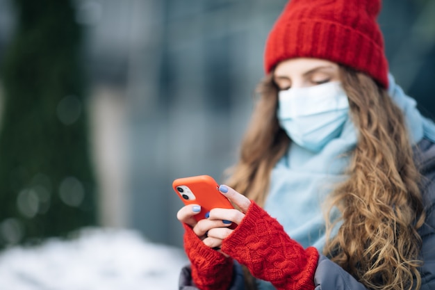 Sluit omhoog portret van mooi Kaukasisch jong krullend haarwijfje in medisch masker texting op mobiel.