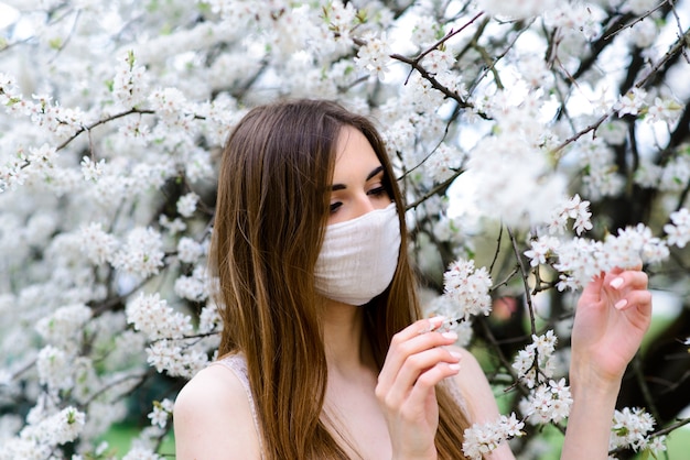 Sluit omhoog portret van teder meisje in een witte blouse onder een bloeiende kersenboom met een masker van het coronavirus.