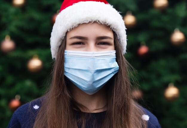 Sluit omhoog Portret van vrouw die een hoed van de Kerstman en medisch masker met emotie draagt. Tegen de achtergrond van een kerstboom. Coronapandemie