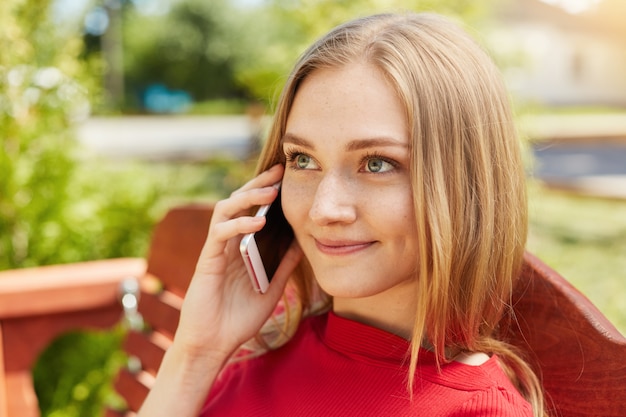 Sluit omhoog portret van vrouw met blauwe ogen met sproeterig gezicht en dunne lippen die bij bank zitten die in openlucht haar vriend over celtelefoon roept. jong wijfje dat over smartphone communiceert die goed humeur heeft