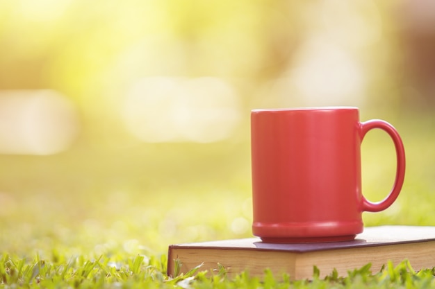 Sluit omhoog rode ceramische koffiekop en oud boek in groene tuin in ochtendtijd