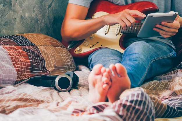 Sluit omhoog van de jonge mens gebruikend tablet en speel elektrische gitaar op zijn bed