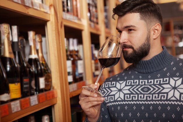 Sluit omhoog van een knappe gebaarde mens die verouderende rode wijn in een glas ruiken, winkelend bij de wijnopslag