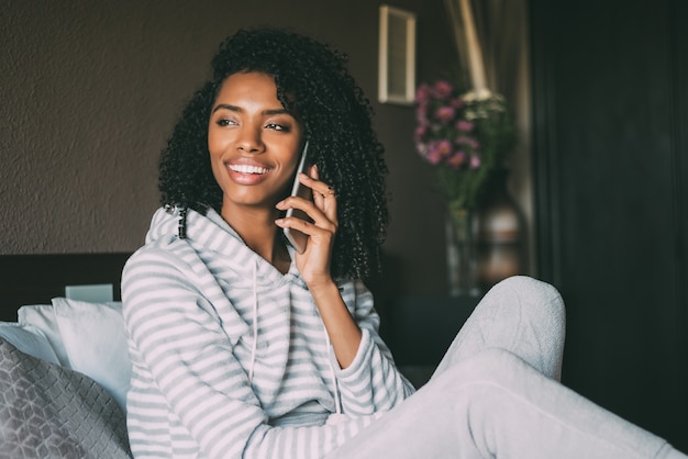 Sluit omhoog van een mooie zwarte die met krullend haar en telefoon op bed glimlacht gebruikt weg kijkend