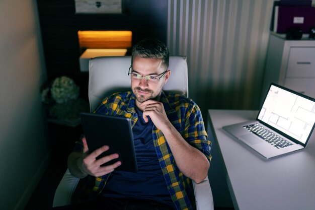 Sluit omhoog van glimlachende ongeschoren werknemer met oogglazen bij het gebruiken van tablet terwijl het zitten in het bureau laat bij nacht