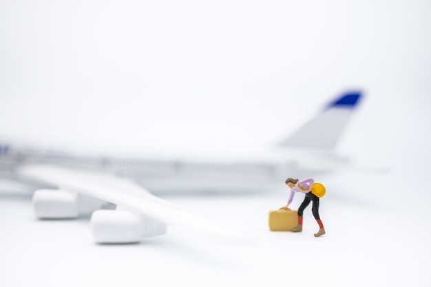 Sluit omhoog van het miniatuurcijfer van de vrouwenreiziger met bagage zich op wit met ministuk speelgoed vliegtuigmodel bevinden.