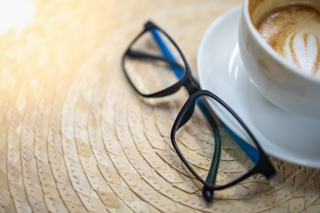 Sluit omhoog van lezingsglazen en witte kop van hete koffie latte met de kunst van de hartvorm op mandewerk.