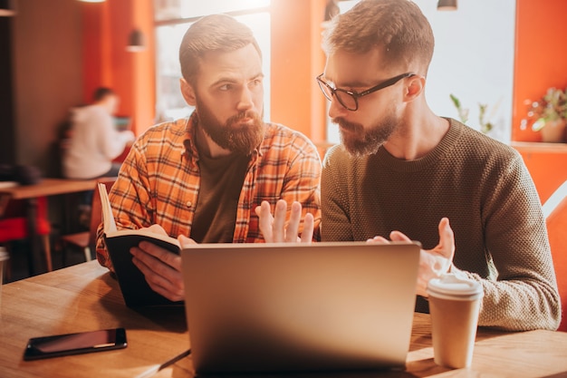 Sluit omhoog van twee gebaarde mannen die samen in een klein koffie zitten. De man aan de rechterkant legt iets uit aan de rechter man die een boek vasthoudt. Ze zien er druk en geconcentreerd uit.