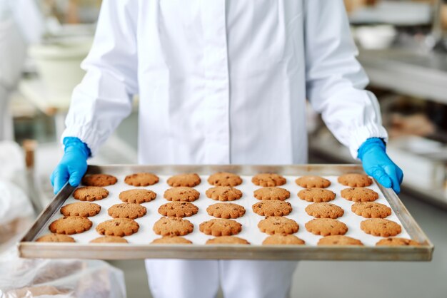 Sluit omhoog van vrouw in steriel eenvormig holdingsdienblad met verse smakelijke koekjes. Voedsel plant interieur.