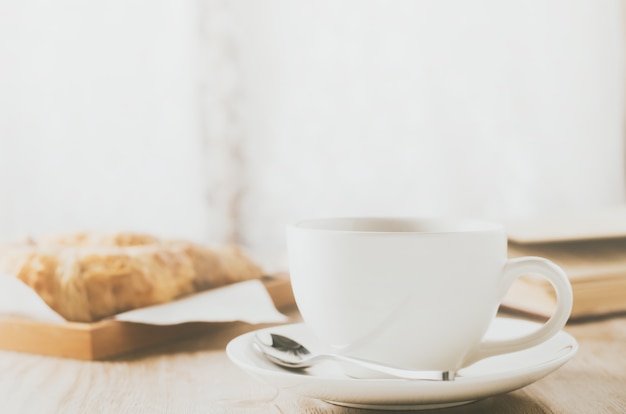 Foto sluit omhoog van witte koffiekop met croissant op houten lijst met uitstekende toon