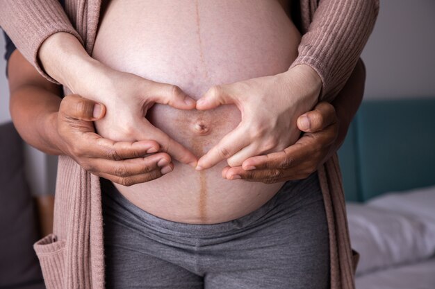 Sluit omhoog van zwangere vrouw die het gebaar van het handhart op buik in slaapkamer maken.