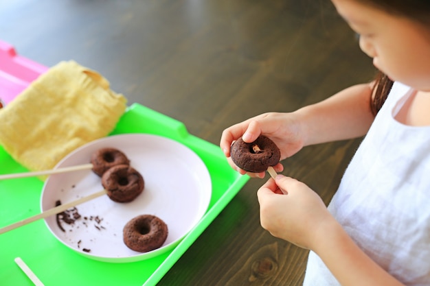 Sluit omhoog weinig Aziatisch jong geitjemeisje in procesklasse van eigengemaakte chocolade donuts.