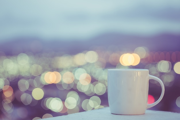 Sluit omhoog witte koffiekop op teller en kleurrijk onduidelijk beeldlicht bokeh van stadsachtergrond