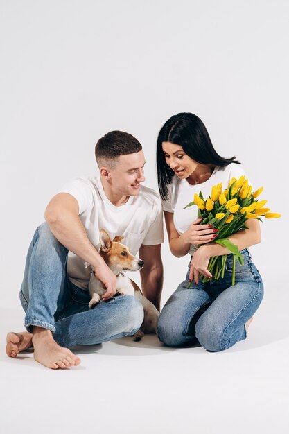 Sluit portret omhoog zitten het jonge paar en koesterend, houdend gele bloemen en hond in studio op witte achtergrond. paar omarmen met dromerige amoureuze uitdrukking. Liefdevolle familie. Vrouwendag vieren.